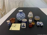 Lot of ceramic collectables including 5 small/miniature vases, a larger blue printed vase, ceramic clogs, a Delft Blue tile, two lidded containers, and a small cup all arranged on a table.