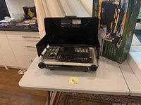 Front view of Coleman InstaStart grill stove on table with box in background, showing two burners, grill plate, control knobs, and side wind guards.
