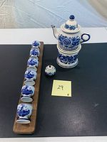Photo of the six Delft Blue ceramic egg cups mounted on a wooden holder, accompanied by a blue and white ceramic teapot on a warmer base, and a small salt container, all on a black table surface.