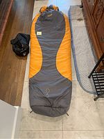 Front exterior of grey and orange mummy sleeping bag laid flat with black carry bag to left