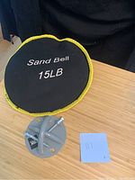 Photo of black 15lb sand bell with yellow trim beside TRX branded X-Mount anchor and screws on wooden surface.