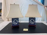Pair of blue porcelain square lamps with foo dog graphics and cream shaded tops, sitting on dark wooden base.