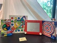 Photo showing Simon game, Blurt game, Etch A Sketch, and tin tambourine together on a black surface with a white tent background and note with item number 49.