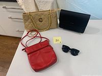 Four items displayed on white table: black Black Nectar sunglasses; red Ili New York leather bag; tan handbag; black Pixie Mood handbag with gold chain straps