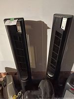 Photo showing two black Honeywell tower heaters/fans side by side. Both have vertical grills and top-mounted control panels with buttons and indicator lights.