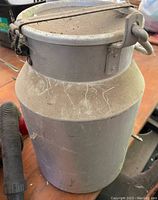 Photo of a single large aluminum milk can with lid and handle, showing signs of heavy use with scratches and dirt.