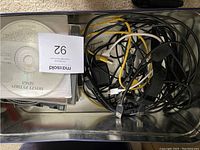 Container filled with miscellaneous cables and CDs including Monty Python Sings.