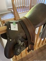 Side view of brown leather English saddle hung on wooden railing, showing seat, flap, and attached stirrup leathers, with some wear marks.