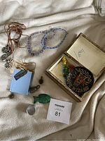 Photo showing two blue/purple beaded necklaces, brown cord necklace with metal pendants, green malachite fish, bronze pilot figurine, blue book-shaped pendant, small magnifying glass, and colorful beaded necklace inside an open box with the voodoo patch.