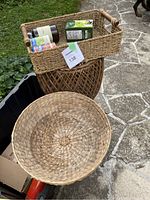 Photo showing three wicker baskets arranged outdoors on stone pavement and black container, including rectangular basket holding three bottles, round open weave basket, and shallow round basket.