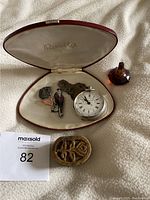 Red leather jewelry box open revealing silver pocket watch, two Art Deco brooches, and gold-backed coral pendant shaped like a shoe; outside the box are a carved sandalwood paste box and a small amber glass bottle.