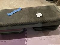 Black step platform on foam mat floor with a blue resistance band on top, showing equipment for aerobic and strength training exercises.