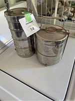 Two stainless steel tiffin lunch cans stacked on a white surface. The left tiffin has four compartments and a rounded handle. The right tiffin has three compartments and a flat, foldable handle.
