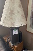 Close-up of one lamp showing dark wooden rectangular base with attached sticker and beige textured lampshade with floral pattern and stitched edges.