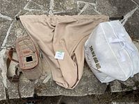 Photo of vintage wicker and leather fishing creel, beige fabric sack, and white fabric bag with gold text laid out on stone surface.