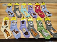 Twelve pairs of women's socks laid out on a wooden table featuring various dog breeds and colors. Each sock depicts a different breed with colorful background and breed names.