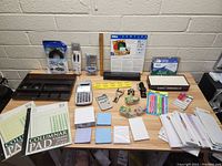 Table showing large assortment of office supplies including pads, calculators, sticky notes, pens, envelopes, tapes, an organizer, and a clipboard.