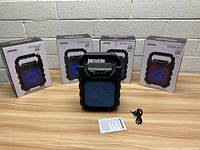 Four boxed Max Power BOOM-M2 LED Bluetooth speakers arranged behind one unpackaged speaker with blue LED light, charging cable, and instruction manual on table