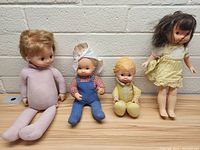Four vintage dolls arranged on a wooden surface with a brick wall background, showing front and side views.