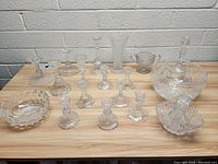 Top view of assorted glass and crystal items including bowls, vases, and candle holders on a wooden table against a white wall.