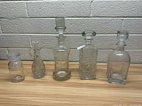 Five assorted glass decanters lined up against a painted brick wall showing their varied shapes and sizes.