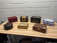 Lot display of seven assorted wooden jewelry boxes on a glass shelf against white brick wall, showing various shapes, sizes and designs