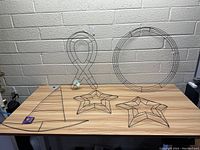 Five assorted metal wire wreath frames positioned on a wooden table against a wall. Shapes visible: circular wreath frame, two star-shaped frames, ribbon awareness loop, and witch hat outline.