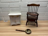 White doll dresser, brown wooden doll chair, vintage bed warmer on floor against white brick wall
