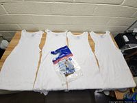 Four white sleeveless men's undershirts laid flat on a table with packaged set of two undershirts included.