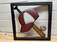 Front view of metal baseball-themed wall hanging inside black square frame on wooden surface with tan brick wall background.