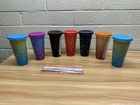 Photo showing seven colorful astrology-themed tumblers with lids on a wooden table against a white brick wall, with a bundled set of straws in front