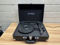 Open briefcase style Victrola record player on a wooden table against a beige brick wall. Shows turntable, tonearm, and control knobs.