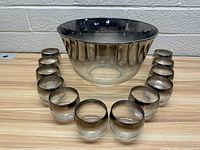 Photo showing large punch bowl with silver rim surrounded by 12 matching small glass cups with silver rims arranged in a semicircle.