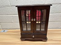 Front view of the wooden jewelry box with two glass doors and a drawer at the bottom, placed against a concrete wall.