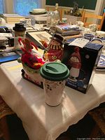 Photo showing the snowman teapot, ceramic bowl with green rim and brown feet, Christmas travel mug with green lid, and boxed Pfaltzgraff Penguin salt shaker on the table among other items.