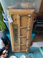 Wooden wall-mounted coat rack with five brass hooks and decorative plaque, angled view showing entire rack and hooks.