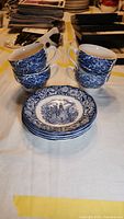 Set of 4 stacked blue and white cups beside 4 stacked matching saucers with colonial scene design on white cloth background