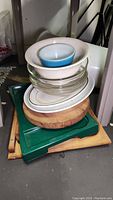 Stack showing multiple items including glass Pyrex baking dishes, white platters, wooden cutting boards, blue and white Pyrex bowl stacked on top of green lap tray.
