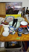 Overview of lot showing small enamel pot, bowls, plates, small pitcher, creamer, utensils and other kitchenware items on table