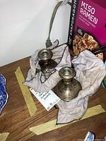 Pair of small Gorham sterling weighted candlesticks wrapped in cloth, showing one candlestick upright and the other lying down, placed on a wood surface near an electrical cord and a box of miso ramen noodles.