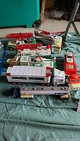 Photo showing multiple toy trucks including three Hess emergency vehicles with ladders, a Hess helicopter, an orange 7 Eleven truck, and other trucks laid out on a cloth.