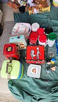 Photo showing a variety of vintage lunch boxes, plastic drink cups, metal tins, and animal-shaped bottle caps arranged on a green fabric surface.