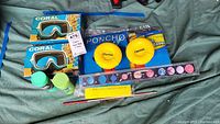 Blue Coral snorkeling masks in boxes, Disney ponchos in packaging, yellow Cheerios holders, space-themed book cover and watercolor paint set with brush on fabric background.
