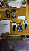 Overview photo showing three Timex wristwatches, Swiss Army trifold wallet in packaging, two hourglass timers, and multicolored bracelet on wooden surface