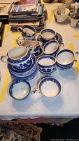 View of multiple blue and white transferware cups and saucers, primarily Old Willow pattern, showing sets grouped on table.
