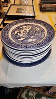 Stack of mostly blue and white transferware dinner plates featuring the Willow Ware pattern with a detailed scenic design and borders.