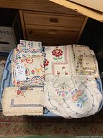 Photo showing assortment of embroidered vintage doilies and napkins on blue plastic tub lid, highlighting various floral and geometric embroidered patterns.