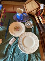 Six white Wedgwood Wellesley dinner plates with embossed floral rim design, four Pfaltzgraff floral plates, blue Bennet Bakeware pitcher and matching blue serving plate, plus a floral cup and saucer on a green cloth.