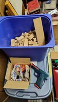 Blue tub filled with assorted natural wood blocks and a box containing a green cap gun toy with cap packs on a gray plastic surface.