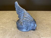 Side view of metal hood ornament showing Indian chief head with feathered headdress and visible wear and texture on base.